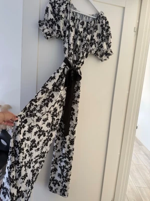 Vit och svart blommig jumpsuit SHEIN - Supersnygg jumpsuit i vitt med svart blommönster och korta puffärmar. Har ett svart midjeband som knyts och vida ben. Perfekt för dig som vill sticka ut med en trendig och bekväm look. Materialet känns lätt och flowy.