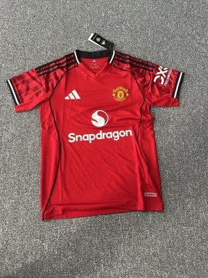 Manchester United matchtröja Adidas - Snygg röd Manchester United matchtröja från Adidas med klubbmärke på bröstet och Snapdragon-logga framtill. Tröjan har svarta och vita detaljer på axlar och ärmslut samt klassiska Adidas-ränder. Tillverkad i lätt och ventilerande funktionsmaterial.
