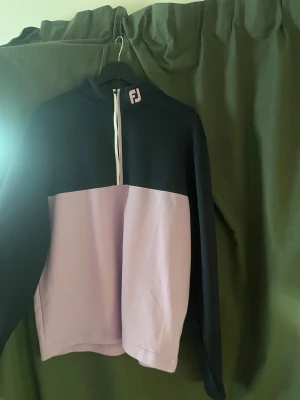 Golftröja FootJoy - Sportig tröja från FootJoy i blått och lila med halv dragkedja framtill och logga på kragen samt ärmen. Tillverkad i mjukt funktionsmaterial, perfekt för golf eller annan träning. Oversized passform och lång ärm för extra komfort.