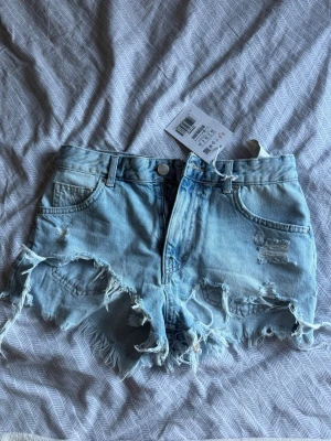 Ljusa denimshorts från Pull&Bear - Säljer ett par ljusblå denimshorts från Pull&Bear med fransig och trasig kant samt slitna detaljer framtill. Klassisk femficksmodell med knapp och dragkedja. Perfekta för varma dagar och en avslappnad stil. Helt oanvända