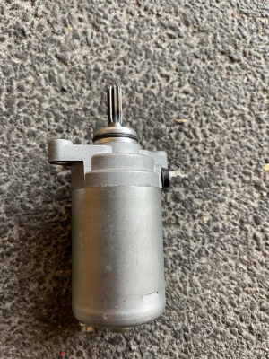Startmotor till moped eller MC - Säljer en startmotor i metall, silverfärgad med cylinderformad kropp och kugghjul i toppen. Passar till moped eller motorcykel. Monteringsfästen och anslutningsskruv synliga på sidan. Perfekt reservdel för dig som gillar att meka.