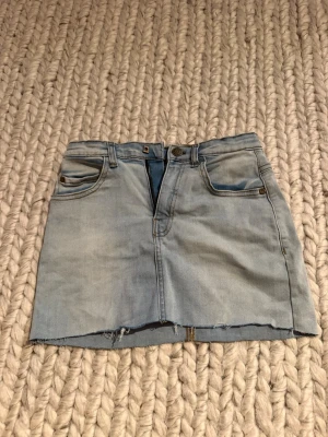 Ljusblå jeanskjol med rå kant - Trendig ljusblå jeanskjol med rå, fransig kant nertill och klassiska fickor fram och bak. Kjolen har bälteshällor och dragkedja med knapp framtill. Perfekt för en avslappnad och cool stil.