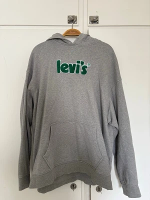 Grå hoodie från Levi's med logga - Säljer en grå hoodie från Levi's i relaxed fit med stor grön logga broderad på bröstet. Hoodien har huva, magficka och ribbade muddar vid ärmslut och nederkant. Perfekt för en chill och avslappnad stil.