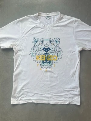 Kenzo t-shirt vit - Säljer en vit t-shirt från Kenzo med ikoniskt tigertryck i blått och gult samt texten 'KENZO PARIS' på bröstet. Klassisk passform och rund halsringning. Perfekt för dig som vill ha en statement tee med streetwear-vibe. Använd få gånger. Hög kvalité. 