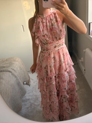 Neo Noir Marli Flower Chiffon Dress - Romantisk långklänning i ljusrosa med blommigt mönster och volanger. Klänningen är ärmlös, har hög hals och fina detaljer vid midjan. Perfekt för sommarens alla tillfällen. Materialet känns lätt och luftigt. Använd en gång under midsommar.