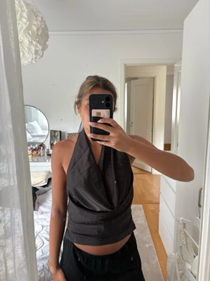 Svart halterneck topp - Snygg svart halterneck topp med draperad framsida och öppen rygg. Toppen har en croppad passform och knyts i nacken. Perfekt för dig som vill ha en trendig och stilren look.