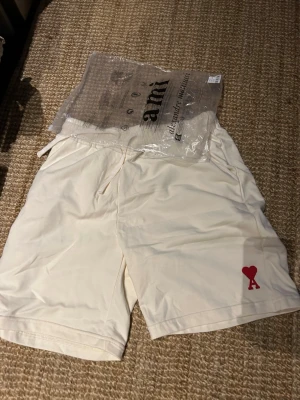 Vita shorts från Ami Paris med hjärtdetalj - Säljer ett par vita shorts från Ami Paris med röd hjärtdetalj och bokstaven A på vänster ben. Shortsen har elastisk midja med snörning och sidofickor. Perfekta för sommaren och riktigt clean look.