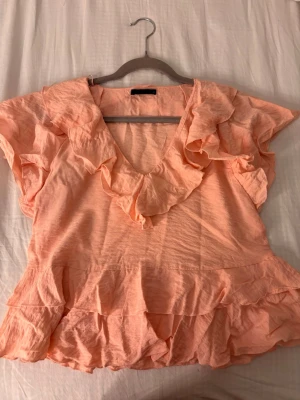 Persikofärgad volangblus från ZARA - Supersöt persikofärgad blus från ZARA med volanger längs ringningen och nederkanten. Perfekt till sommaren! Den har ett litet hål som man ser på bilden. Hålet går däremot att täcka med volangen vid ringningen. 