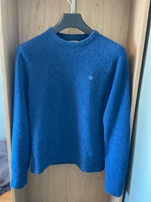 Blå stickad tröja från J.Lindeberg - Snygg blå stickad tröja från J.Lindeberg med rund halsringning och diskret logga på bröstet. Tröjan har en mjuk, lite fluffig struktur och långa ärmar. 