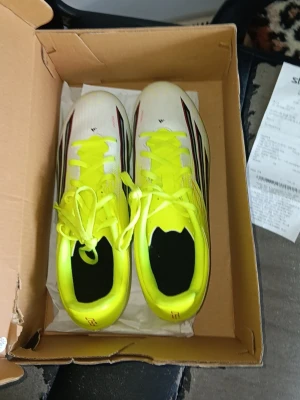 Adidas fotbollsskor gul/vit - Säljer ett par Adidas fotbollsskor med en snygg design i neon-gult och vitt, med svarta och röda detaljer på sidan. Skorna har klassiska snörningar och är tillverkade i syntetmaterial som ger bra grepp och komfort på planen. Perfekta för dig som vill sticka ut på matchen.