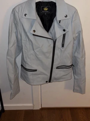 Ljusgrå skinnjacka från Route 66 - Snygg ljusgrå skinnjacka från Route 66 med asymmetrisk dragkedja, tre framfickor med svarta dragkedjor och coola detaljer vid axlarna. Jackan har klassisk bikerstil och krage med tryckknappar. Perfekt för dig som vill ha en stilren och edgy look.