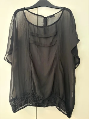 Svart transparent blus från Vero Moda strl.XL - Snygg svart blus från Vero Moda i transparent, lätt material. Blusen har rund halsringning, korta ärmar och dekorativa lager framtill. Nederkanten är smockad för en avslappnad look. Perfekt att styla med jeans eller kjol.
