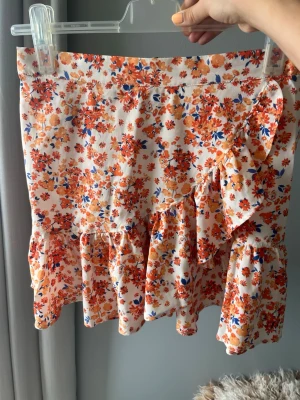 Blommig volangkjol med omlott - Kort kjol med volang och omlottdetalj i vitt tyg med orange, röda och blå blommor. Kjolen har resår i midjan och är perfekt för sommarens alla tillfällen. Lätt och luftig modell som ger en söt och trendig look.