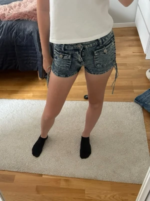 Blå  denimshorts med snörning - Snygga grå denimshorts med  snörning i sidorna. Shortsen har två framfickor och knappdetaljer samt en lågmidjad passform. Perfekta för sommardagar och ger en avslappnad vibe.