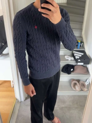 Morris Mörkblå kabelstickad tröja  - Snygg mörkblå kabelstickad tröja från Morris med rund halsringning och röd broderad logga på bröstet. Ny pris 1 700 kr, lite urtvättad men annars bra skick. 