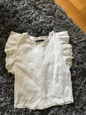 Vit ribbad topp med volangärmar Zara - Säljer en vit ribbad topp från Zara med korta volangärmar. Toppen har en rund halsringning och är i ett mjukt, stretchigt material som ger en bekväm passform. Perfekt för att lyfta vilken outfit som helst med sina snygga detaljer.