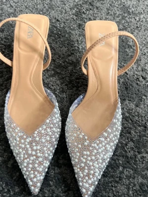  pumps från Zara - Säljer ett par eleganta pumps från Zara med spetsig tå och dekorerade med vita pärlor och glittrande stenar. Skorna har en beige innersula och smala remmar runt hälen. Perfekta för dig som vill sticka ut med extra bling.