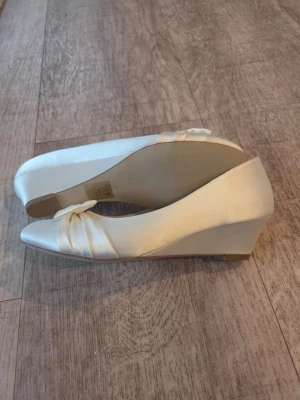 Satinskor med kilklack och rosett - Snygga ballerinaskor med kilklack i färgen Ivory och  rosett framtill. Skorna har rundad tå och är tillverkade i ett lyxigt satintyg som ger en elegant look. Perfekta för dig som brudsko, fest eller student/examensko