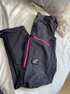 Svarta täckbyxor från Rymac - Snygga svarta friluftsbyxor från Revolution Race med rosa dragkedja på benfickan och elastisk midja. Byxorna har praktiska fickor och förstärkta partier. Materialet är slitstarkt och vattenavvisande. Pruta om så behövs, vill bara bli av med de😄