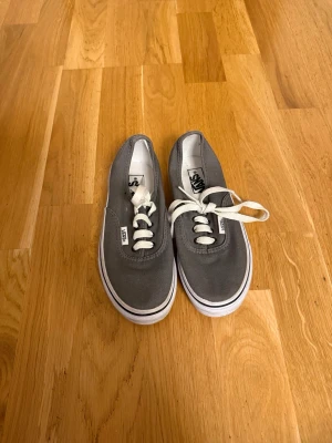 Gråa Vans låga sneakers - Säljer ett par klassiska gråa Vans sneakers med vita snören och vit sula. Skorna har den ikoniska Vans-loggan bak på hälen och är tillverkade i canvasmaterial. Perfekta för en avslappnad och stilren look.