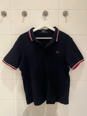 Mörkblå piké från Fred Perry - Snygg mörkblå pikétröja från Fred Perry med klassisk krage och knappar. Detaljer i vitt och rött på krage och ärmslut samt den ikoniska loggan broderad på bröstet. Perfekt till jeans eller shorts för en sportig look.