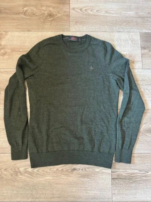 Olivgrön stickad tröja Morris  - Snygg olivgrön stickad tröja från morris med rund halsringning och diskret broderad logga på bröstet. Tröjan har långa ärmar och ribbade muddar vid ärmslut och nederkant. Perfekt för lager på lager och enkel att matcha.