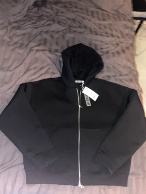 Scooba dubble zip up hoodie från Weekday - Svart hoodie från Weekday i storlek L med huva och hel dragkedja framtill. Tröjan har en clean look, stora fickor och ribbade muddar. Materialet är mjuk bomull blandat med polyester, perfekt för chill dagar. Snygg och enkel att matcha med jeans eller joggers.