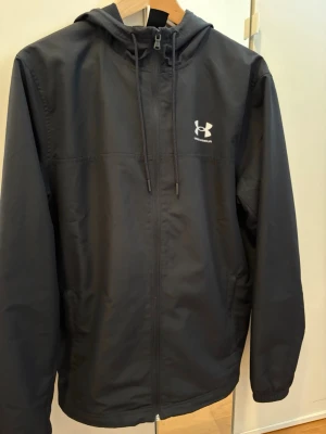 Svart vindjacka från Under Armour - Snygg svart vindjacka från Under Armour med huva och dragkedja framtill. perfekt för träning eller vardagligt bruk. Tillverkad i lätt och vindtätt material, perfekt för blåsiga dagar. 