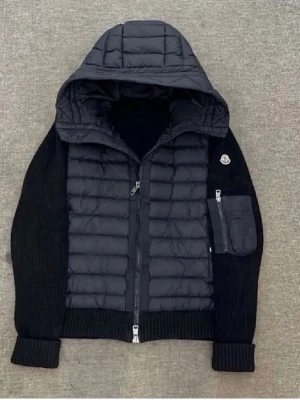 Marinblå pufferjacka med huva från Moncler - Snygg marinblå pufferjacka från Moncler med vadderad front och huva. Jackan har stickade ärmar och ribbade muddar, samt en praktisk ficka med dragkedja på ena ärmen. Perfekt för kalla dagar och riktigt trendig look. ❗️upplevs svart men är marinblå❗️ pris kan diskuteras 🤩skriv vid frågor!