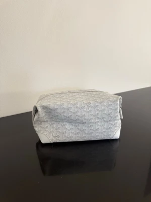 Vit necessär från Goy - Snygg vit necessär från Goy med klassiskt mönster och dragkedja upptill. Väskan har en boxig form och detaljer i vitt läder. Perfekt för att organisera småprylar eller smink i väskan.