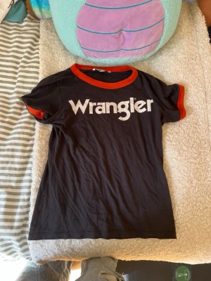 Svart Wrangler t-shirt med röd kant - Säljer en svart t-shirt från Wrangler med vit logga framtill. T-shirten har röda kanter vid halsringning och ärmslut, vilket ger en retrovibe. Tillverkad i mjuk bomull och har en klassisk passform med korta ärmar. 