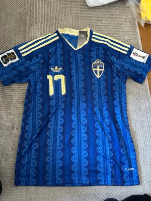 Blå Sverige matchtröja Adidas - Säljer en blå matchtröja från Adidas med Sveriges landslagsemblem och nummer 17 i gult. Tröjan har retroinspirerat mönster, tre gula ränder på axlarna och V-ringad krage. Tillverkad i lätt och ventilerande material, perfekt för fotboll eller träning.