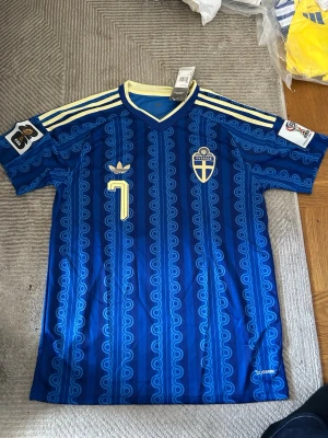 Blå Sverige fotbollströja Adidas - Säljer en blå Sverige fotbollströja från Adidas med retroinspirerat mönster, gula detaljer och nummer 7 på bröstet. Tröjan har broderat landslagsemblem, Adidas-logga och tre gula ränder på axlarna. Perfekt för dig som älskar fotboll och vill sticka ut på läktaren.