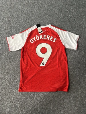 Arsenal matchtröja Gyökeres #9 - Röd och vit Arsenal matchtröja från Adidas med Gyökeres och nummer 9 på ryggen. Tröjan har klubbmärke, sponsorlogga och klassiska Adidas-ränder på axlarna. Tillverkad i lätt och ventilerande funktionsmaterial, perfekt för fotbollsträning eller match.