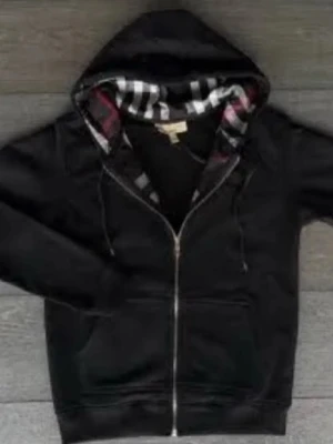 Svart hoodie från Burberry med dragkedja - Säljer en svart hoodie från Burberry med dragkedja och klassiskt rutigt foder i huvan. Hoodien har två fickor framtill, justerbar dragsko i huvan och är tillverkad i mjukt material. Perfekt för dig som gillar stilrena plagg med exklusiv känsla.