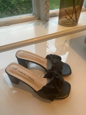 Svarta klackade sandaler - Säljer ett par svarta sandaler med breda remmar i glansigt material och fyrkantig tå. Skorna har en chunky klack som ger höjd och en trendig look. Perfekta för dig som vill ha något stilrent men ändå edgy. Endast testade!