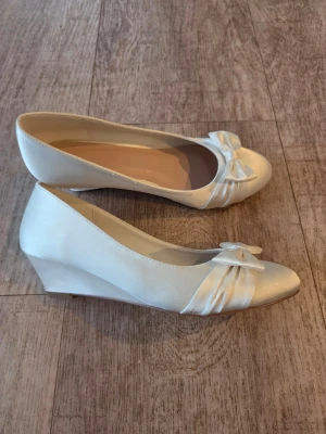 Satinskor med kilklack och rosett st 38  - Snygga Satinskor med kilklack och rosett framtill. Färg Ivory Skorna har rund tå och är tillverkade i ett lyxigtt satin som ger en elegant look. Perfekta för dig som vill ha något stilrent men ändå lite extra.