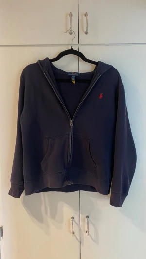 Mörkblå hoodie från Polo Ralph Lauren - Mörkblå hoodie med dragkedja från Polo Ralph Lauren. Klassisk design med röd broderad logga på bröstet, känguruficka framtill och huva. Perfekt för en chill och avslappnad stil. Mjuk bomullskänsla och ribbade muddar vid ärmslut och nederkant. Barn stolen L som motsvar Xs