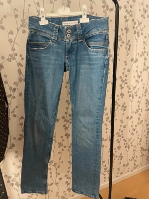 Blåa jeans med dubbla knappar - Säljer ett par ljusblå jeans från pepe jeans med raka ben och dubbla knappar i midjan. Jeansen har klassiska fem fickor och kontrastsömmar. Tillverkade i stretchig denim för skön passform. Storlek 27 som är som en s ungefär 