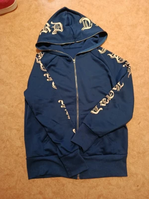 Blå hoodie med tryck och dragkedja - Snygg blå hoodie med dragkedja och huva. Hoodien har vita tryck i gotisk stil på huvan och längs båda ärmarna. Muddar vid ärmslut och nederkant. Perfekt för en streetwear-look.