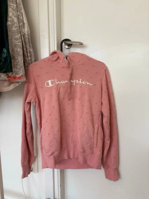 Rosa hoodie från Champion - Säljer en rosa hoodie från Champion med vit logga framtill och små mönstrade C-detaljer över hela tröjan. Hoodien har huva, långa ärmar och ribbade muddar. 
