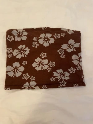 Brun blommig tubtopp från H&M - Säljer en brun tubtopp från H&M med vita blommor i ett somrigt mönster. Toppen är axelbandslös och har en enkel, rak passform. Perfekt för varma dagar och enkel att matcha med jeans eller kjol.