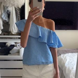 Blå oneshoulder topp med volang - Säljer en ljusblå oneshoulder topp med stor volangdetalj över axeln. Toppen har en strukturerad yta och är perfekt för dig som vill sticka ut lite extra. Passar snyggt till både jeans och kjol. Från Lindex i stl xs, toppen skick! 