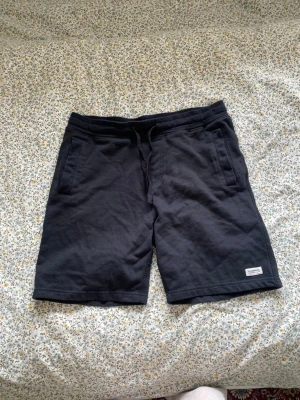 Svarta mjukisshorts med snörning - Svarta mjukisshorts i bomull med snörning i midjan och två sidofickor samt en bakficka. Perfekta för chill dagar eller träning. Liten patch med logga på vänster ben.