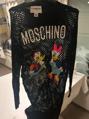 Svart nättröja Moschino Disney - Cool svart långärmad nättröja från Moschino med stort tryck av Kalle Anka och Kajsa Anka på framsidan. Oversized passform och stora vita bokstäver med Moschino-logga upptill. Perfekt statement-plagg för dig som gillar unika designer.