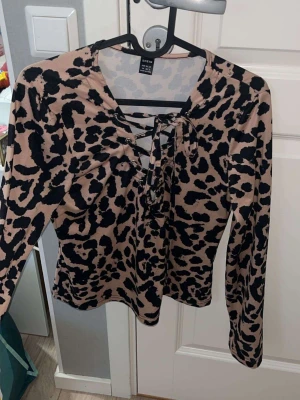 Leopardmönstrad topp från SHEIN - Snygg leopardmönstrad topp från SHEIN i beige och svart. Toppen har långa ärmar, snörning framtill och en v-ringad hals. Tillverkad i ett mjukt och stretchigt material som sitter bekvämt på kroppen.