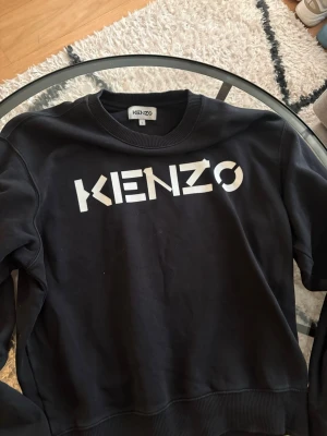 Svart sweatshirt Kenzo - Säljer en svart sweatshirt från Kenzo med stor vit logga framtill. Tröjan har rund halsringning, ribbade muddar vid ärmslut och nederkant samt en avslappnad passform. Perfekt för dig som gillar streetwear och vill ha en stilren look.
