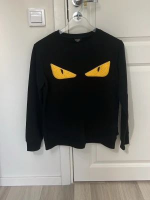 Fen och di  - Säljer en svart sweatshirt från Fen och di med ikoniska gula ögon framtill. Tröjan har rund halsringning, långa ärmar och ribbade muddar. Tillverkad i mjukt material som känns skönt mot huden. Perfekt för dig som vill sticka ut med en cool och unik design.