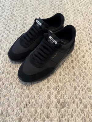 Axel Arigato Genesis  - Snygga svarta sneakers från Axel Arigato med klassisk låg siluett. Skorna har ovandel i mocka och textil, svarta skosnören och diskret logga i guld på sidan. Perfekta för en clean och stilren look.