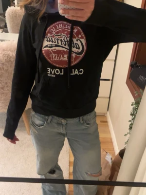 Svart hoodie California Team - Svart hoodie med tryck i vitt och vinrött från California Team. Stor rund logga framtill och texten 'CAL LOVE'. Huva med dragsko och magficka. Perfekt för en avslappnad streetstil.
